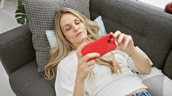 Sarışın kadın, modern oturma odasındaki akıllı telefonuyla kanepede uzanıyor.