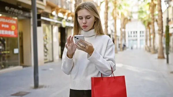 A blonde woman with blue eyes using a smartphone outdoors in a city street, carrying a shopping bag.