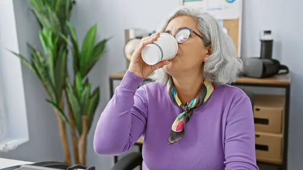 成熟した女性は カジュアルなプロの休憩の瞬間を反映して 近代的なオフィスの設定でコーヒーを飲む — ストック写真