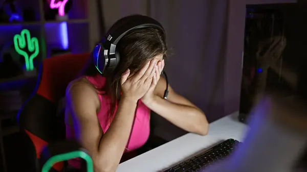 Exhausted young, beautiful hispanic woman streamer stressed from overworked gaming stream at dark gaming room, unhappy portrait of tired female with attractive hairstyle playing on computer