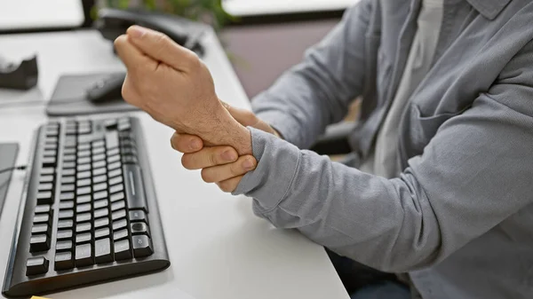 A hispanic man in an office wears a grey shirt, holding his wrist, denoting possible pain or injury, next to a computer keyboard.