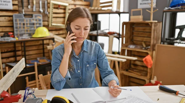 A focused woman multitasking in a carpentry workshop while on a phone call and writing notes.