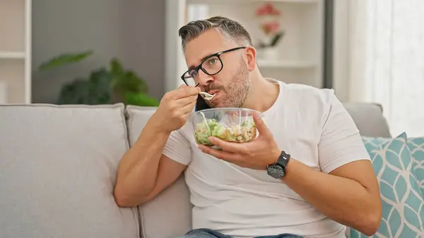 Grey-haired man eating salad talking on smartphone at home