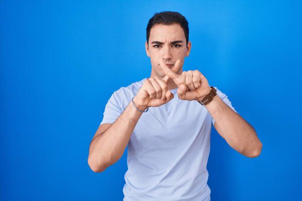 Young hispanic man standing over blue background rejection expression crossing fingers doing negative sign 