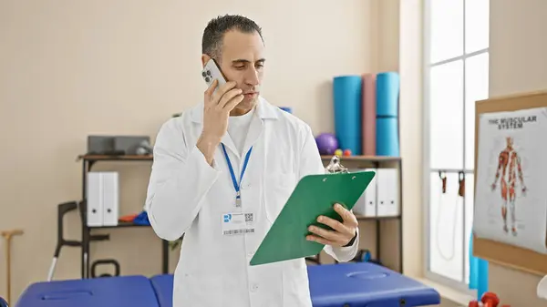 Hispanic pharmacist in lab coat talking on phone and holding clipboard in clinic.
