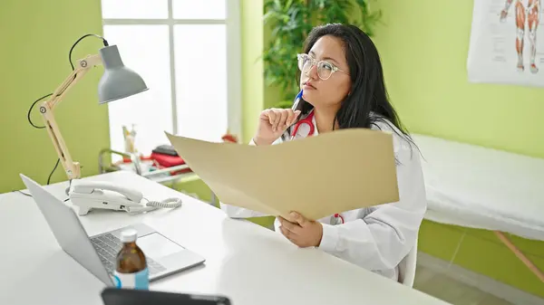 Young chinese woman doctor reading medical report thinking at the clinic