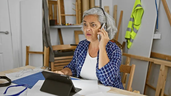 Mature woman on phone multitasking in workshop with tablet and blueprints.