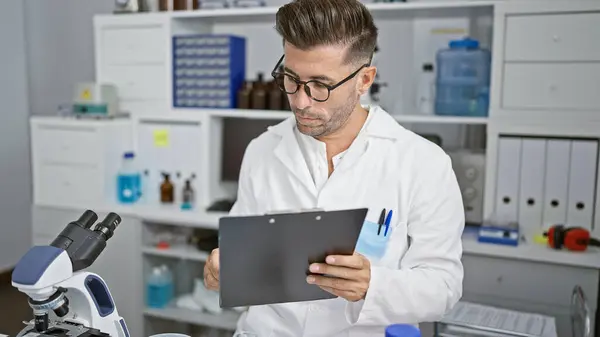 Handsome young hispanic man, charismatic scientist engrossed working, writing a research report in bustling chemistry lab, using a microscope and test tubes for a crucial medical experiment.