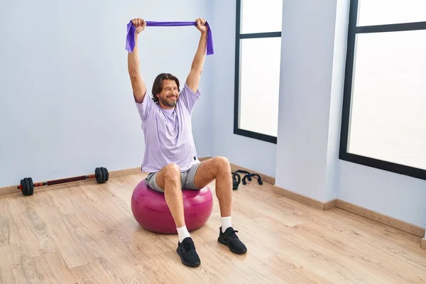 Middle age man training with elastic band sitting on fit ball at sport center