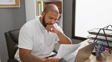 Odaklanmış Afro-Amerikan bir adam ofis masasında dizüstü bilgisayar ve malzemelerle evrak okuyor..