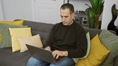 Modern bir oturma odasında dizüstü bilgisayarla koltukta oturan neşeli bir adam..