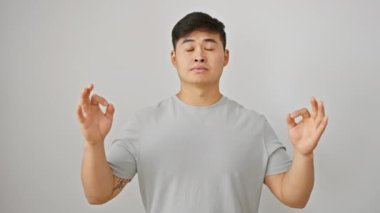 Zen 'le uyum içinde, genç Asyalı adam yoga pozu verir. Tişört giyer, sakin olur, rahatlar, izole edilmiş beyaz arka planda gözlerini kapatır.
