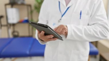 Beyaz önlüklü orta yaşlı bir adam klinik ortamda tablet kullanıyor. Bu da sağlık sektöründe profesyonellik olduğunu gösteriyor..