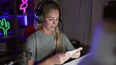 Sarı saçlı ve kulaklıklı odaklanmış genç bir kadın geceleri neon ışıklı bir oyun odasında akıllı telefon kullanıyor..