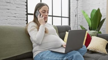 Hamile bir İspanyol kadın evde oturuyor, dizüstü bilgisayar kullanıyor ve akıllı bir telefonla konuşuyor, bağlantı ve annelik temalarını çağrıştırıyor..