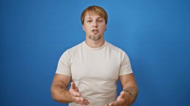 Sarışın, sakallı ve dövmeli yakışıklı bir genç açık havada mavi bir duvara poz veriyor.