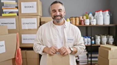 Gri saçlı, gönüllü tişörtlü, sırıtan sakallı bir adam, bir depoda duruyor, üzerinde 