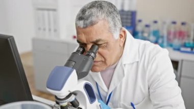 Laboratuvar önlüğü giymiş olgun bir adam laboratuvar ortamında mikroskop kullanırken gülümser..