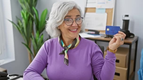 一个快乐的中年白发女人 在办公室里笑着 拿着信用卡时竖起了一个凉爽的大拇指 欢呼成功的理财和自信的生活方式 — 图库视频影像