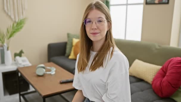一位面带微笑的高加索女人 金发碧眼 戴着眼镜 坐在一间有沙发和装饰植物的现代客厅里 — 图库视频影像