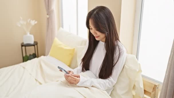 Asiatin Mit Smartphone Einem Modernen Sonnendurchfluteten Schlafzimmer Mit Weißer Bettwäsche — Stockvideo