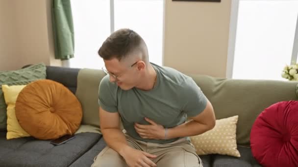 若いヒスパニック男性は 家庭の設定でソファーに座っている間 胃痛を経験します — ストック動画