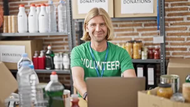 バックグラウンドの物資の棚を持つ寄付センターの倉庫でラップトップで作業するボランティアのシャツを着て笑う金髪の男 — ストック動画