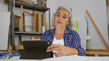Marangozluk araçlarıyla çevrili ve konsantrasyon ve yaratıcılığı yansıtan atölyede tablet kullanan olgun bir kadın.