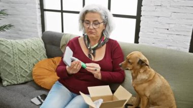 Gözlüklü olgun bir kadın, internet alımından kafası karışmış, akıllı telefondan köpek eşliğinde oturma odasında yardım istiyor..