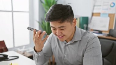 Ofisteki Asyalı adam akıllı telefon kullanırken gülümsüyor ve sıradan bir iş ortamı yaratıyor..