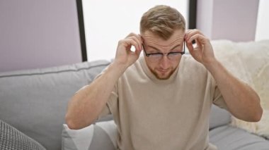 Gri bir koltukta oturan sakallı, genç ve yakışıklı bir adam kırmızı bir kitap okuyor..