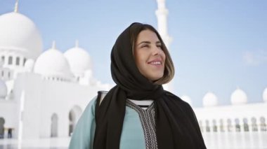 Başörtüsü takan gülümseyen bir kadın Abu Dabi 'deki beyaz kubbeli büyük caminin önünde duruyor..