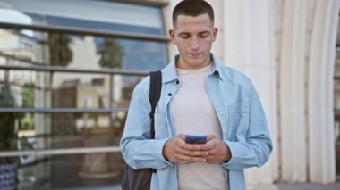 Sırt çantalı genç bir adam modern cam binanın önünde telefonuna mesaj atıyor..