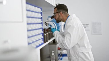 Laboratuvar ortamında mikroskopla çalışan sakallı genç bir erkek bilim adamı profesyonellik ve detaylara dikkat çekiyor..