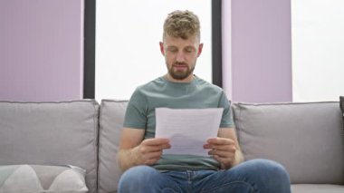 Genç, sakallı bir adam, modern, iyi ışıklandırılmış bir oturma odasında, rahat ve odaklanmış bir atmosfer yayarak bir kanepede otururken bir belge okur..