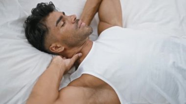 Beyaz tişörtlü, gevşemiş bir İspanyol adam yatağında uzanıyor. Huzur ve rahatlığı bir ev ortamında somutlaştırıyor..