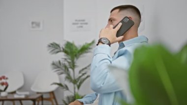 Lobide akıllı telefondan konuşan İspanyol bir adam saat, kapalı alanda yeşillik ve sandalyelerle konuşuyor..