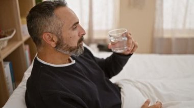 Gri saçlı, olgun sakallı bir adam evdeki aydınlık bir yatak odasında dinlenirken su içer. Huzur ve özgüven hissi uyandırır..