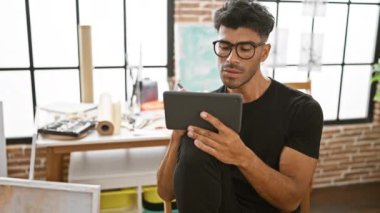 Gözlüklü ve sakallı bir İspanyol iyi ışıklandırılmış bir stüdyoda tablet kullanıyor..
