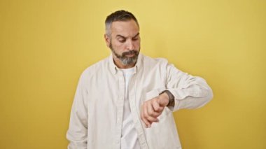 Beyaz tişörtlü olgun sakallı adam sarı arka planda şaşkınlık ve endişe dolu bir kontrol süresini ifade ediyor..