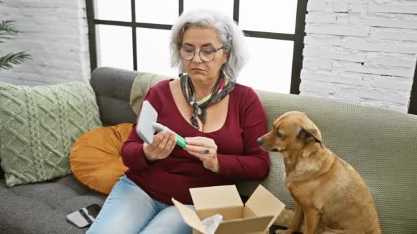 オンライン購入に裏打ちされたメガネ付き成熟した女性は居心地の良いリビングルームで犬の伴侶とスマートフォンで支援を求めます — ストック動画