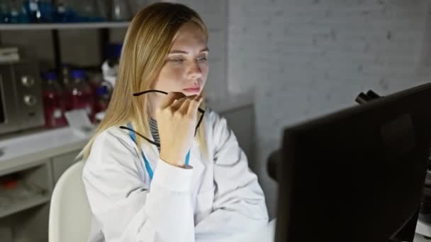 メガネをかけたブロンドの女性が 現代の実験室でコンピュータを意図的に使っている — ストック動画