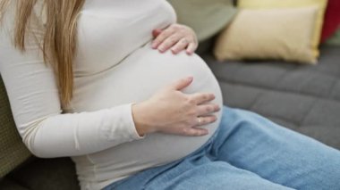 Huzurlu genç bir kadın karnını nazikçe kucaklar, rahat bir oturma odasında beklenti ve neşe saçar..