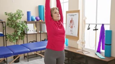 Bir kadın rehabilitasyon kliniğinde egzersiz yapıyor, iskelet modelinin ve terapi ekipmanlarının önünde geriniyor..