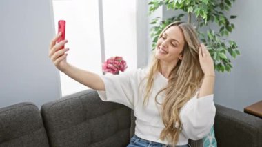 Genç bir kadın, modern bir oturma odasında, akıllı telefonlu bir selfie çekiyor, rahat bir ev atmosferi ve gündelik tarzını sergiliyor..