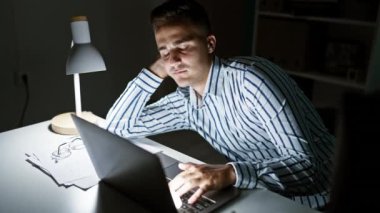 Karanlık bir ofis odasında dizüstü bilgisayar kullanırken esnemekten yorgun düşmüş bir adam..