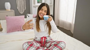 Pijamalı, gülümseyen, mavi bardağı parlak bir yatak odasında tutarken telefonuyla selfie çeken İspanyol bir kadın..