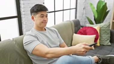 Genç Asyalı bir adam, modern bir oturma odasında elinde uzaktan kumandayla kanepede uzanıyor..