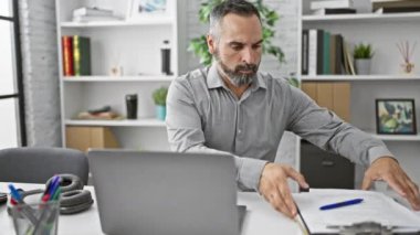 Ofisteki olgun sakallı bir adam gömleğini dizüstü bilgisayarla, belgelerle ve modern dekorla düzeltiyor..