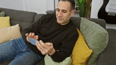Gülümseyen İspanyol adam rahat modern oturma odasında akıllı telefon kullanıyor.